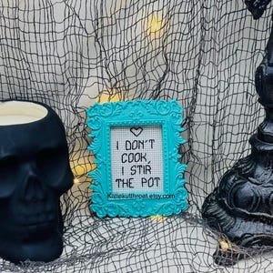 May include: A small, rectangular, bright turquoise ornate frame containing a white cross-stitch piece with a black heart icon and the text: I DON'T COOK, I STIR THE POT. Below the text is the URL: Katiekutthroat.etsy.com in black lettering.