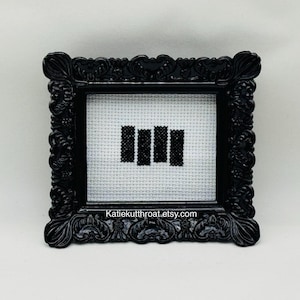 May include: A small, rectangular black ornate frame containing a white Aida cloth with a black cross-stitched design of four vertical bars. The text Katiekutthroat.etsy.com is printed in white at the bottom of the frame's inner border area.