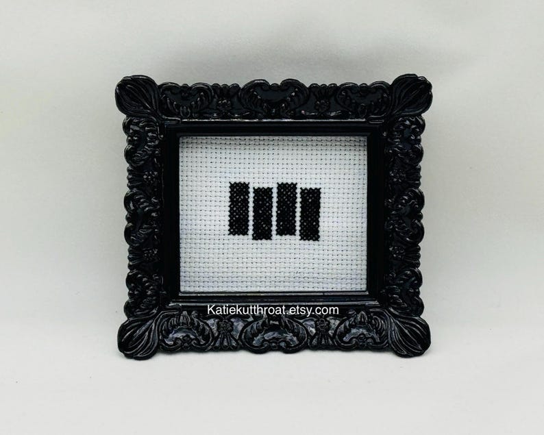 May include: A small, rectangular black ornate frame containing a white Aida cloth with a black cross-stitched design of four vertical bars. The text Katiekutthroat.etsy.com is printed in white at the bottom of the frame's inner border area.