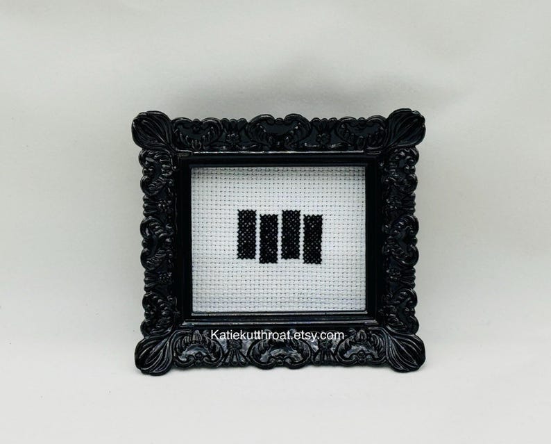 May include: A small, square, black ornate frame containing a cross-stitch piece on white Aida cloth. The embroidery features four vertical black bars arranged side-by-side. The text 'Katiekutthroat.etsy.com' is printed at the bottom of the frame.