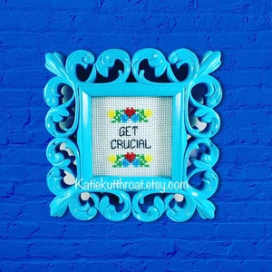 May include: A blue framed cross-stitch with the text "Get Crucial" in white thread. The cross-stitch is framed in a light blue ornate frame and is hanging on a blue brick wall.