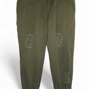 Puede incluir: Pantalones cargo verde oliva con cintura con cordón, bolsillos y parches desgastados. Los pantalones están colgados de una percha negra con pinzas de madera.