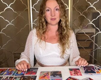 Könnte beinhalten: Eine Frau sitzt an einem Tisch mit ausgebreiteten Tarotkarten. Die Karten zeigen farbenfrohe Illustrationen und Text. Die Frau trägt ein weißes Oberteil. Der Hintergrund umfasst einen dekorativen Bildschirm mit Mond- und Halbmond-Design.