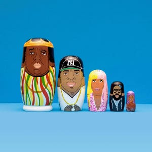 May include: A set of five nesting dolls, each depicting a different person. The largest doll is a black man with a gold crown, the next is a black man wearing a baseball cap with "NY" on it, the third is a woman with blonde hair, the fourth is a black man with a beard, and the smallest is a black woman with purple hair.