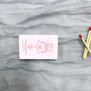 May include: A white matchbox with a red illustration of a birthday cake with candles and the text "Angel's Birthday 05.03.25 123 Anywhere St, Any City"