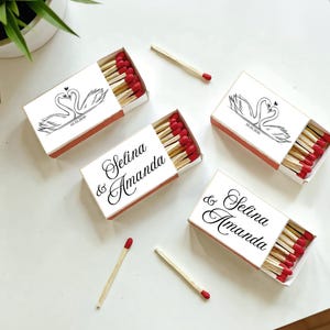 May include: White matchboxes with red-tipped matches. The boxes feature a black swan design and the names "Selina & Amanda" in elegant script. A few loose matches are scattered around the boxes, creating a wedding theme.