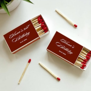 May include: Two rectangular matchboxes with dark red covers featuring white cursive text that reads Anna's 21st Birthday and Anna's Birthday with the date 04.07.2026. Each box contains wooden matches with red tips, and two loose matches lie nearby.