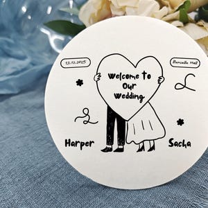 Pode incluir: Uma placa de casamento redonda e branca com uma ilustração a preto e branco de um casal segurando um coração com a frase "welcome to our wedding". Os nomes "Harper" e "Sacha" estão abaixo.