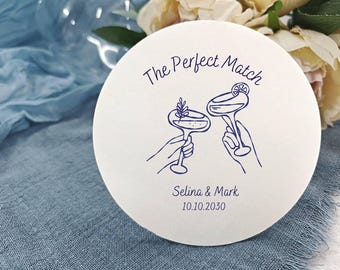 Può includere: Un sottobicchiere rotondo bianco sporco con la scritta "The Perfect Match" e due bicchieri da cocktail in inchiostro blu. Sotto, i nomi "Selina & Mark" e la data "10.10.2030".