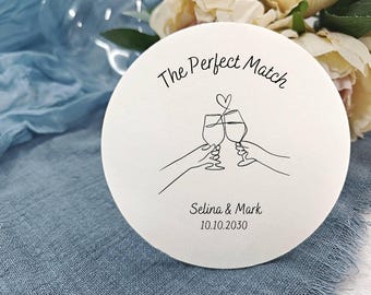Può includere: Un sottobicchiere rotondo bianco con la scritta "The Perfect Match" e un disegno a tratto di due calici di vino che si toccano. Sotto il disegno ci sono i nomi "Selina & Mark" e la data "10.10.2030".