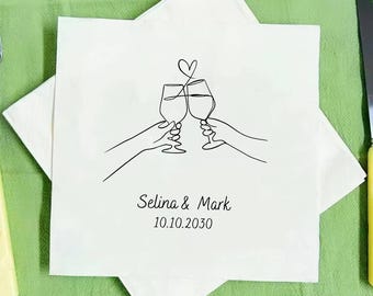 May include: White square napkin with a black line drawing of two hands toasting wine glasses, topped with a heart. The names "Selina & Mark" and the date "10.10.2030" are printed below the illustration. The napkin is on a green surface.