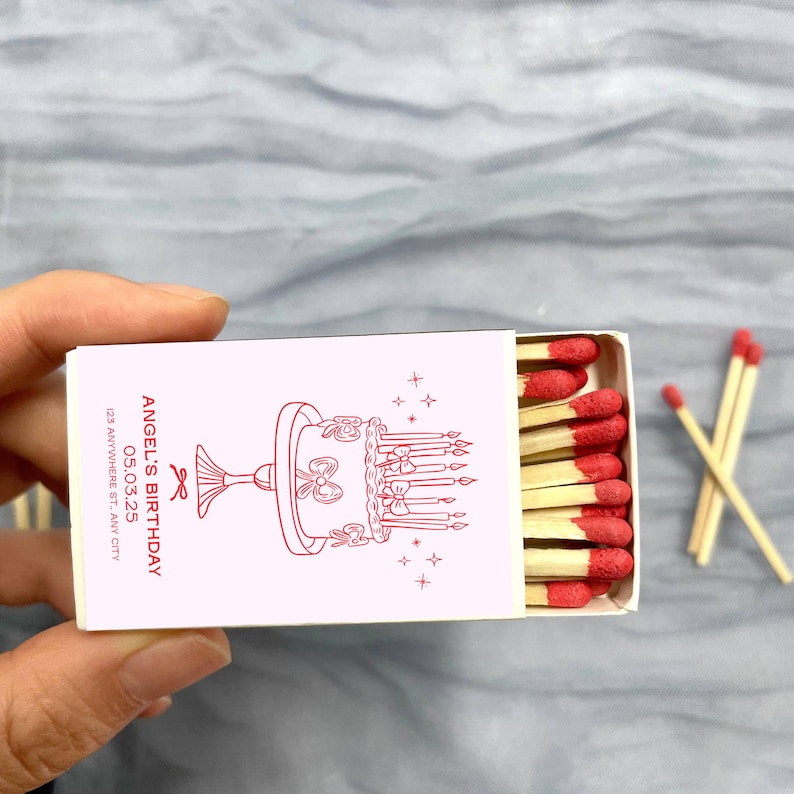 May include: A white matchbox with a pink label. The label has a red illustration of a birthday cake with candles and the text "ANGEL'S BIRTHDAY 05.03.25 123 ANYWHERE ST, ANY CITY". The matchbox is open and contains red-tipped matches.