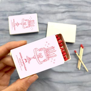 May include: A pink matchbox with a red illustration of a birthday cake with candles and a ribbon. The matchbox has the text "Angel's Birthday 05.03.25 123 Anywhere St, Any City" printed on it.