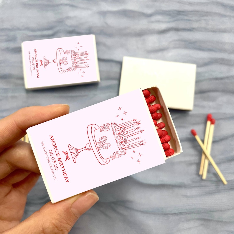 May include: A pink matchbox with a red illustration of a birthday cake with candles and a ribbon. The matchbox has the text "Angel's Birthday 05.03.25 123 Anywhere St, Any City" printed on it.