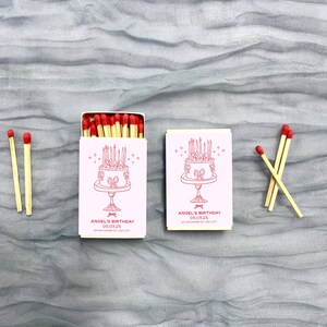 May include: Two pink matchboxes with a white cake design and the text "Angel's Birthday 03.03.25" on the front. Each box contains matches with red heads.