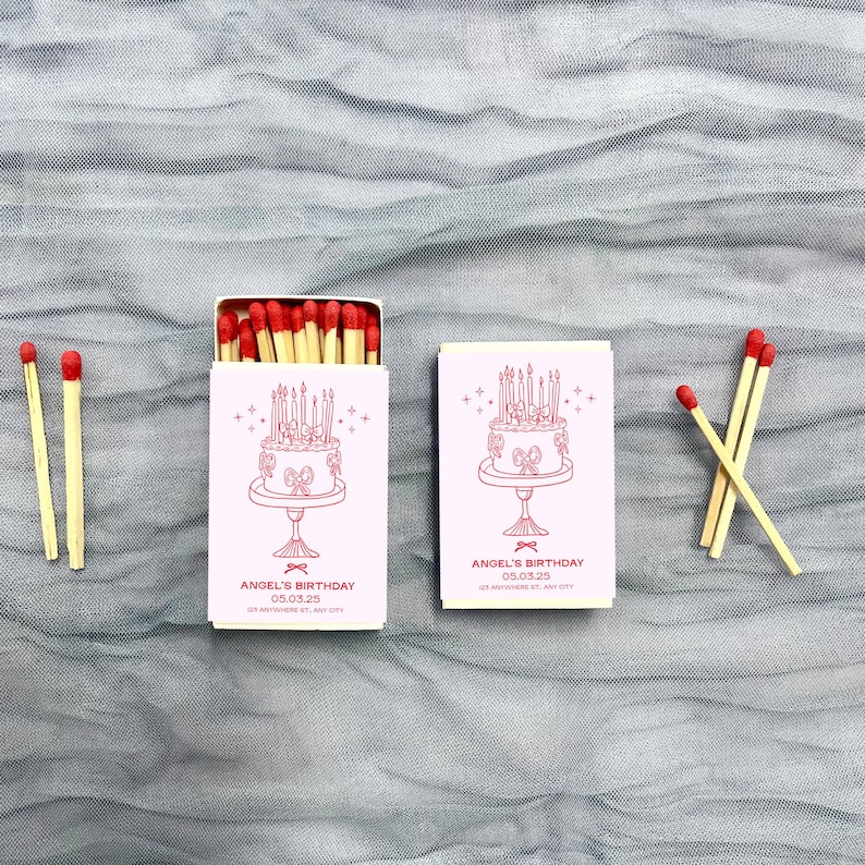May include: Two pink matchboxes with a white cake design and the text "Angel's Birthday 03.03.25" on the front. Each box contains matches with red heads.