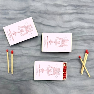 May include: Three matchboxes with pink and white labels. The labels have a red illustration of a cake with candles and the text "Angel's Birthday 03.03.23".