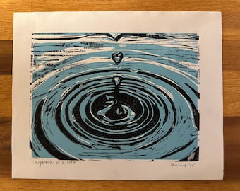May include: A black and blue linocut print depicting a heart-shaped droplet falling into water, creating concentric circles. The print includes the text "compassion is a verb".