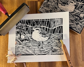 May include: A black and white linocut print of a bird standing in a marshy area. The print is on white paper, with a wooden block and carving tools nearby. A red and black brayer is also visible.