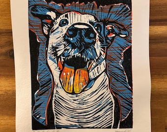 May include: A linocut print of a dog with a happy expression. The dog's face is white with black, blue, and orange accents. The dog's tongue is orange, and the print is on white paper. The name "Roscoe" is printed at the bottom.