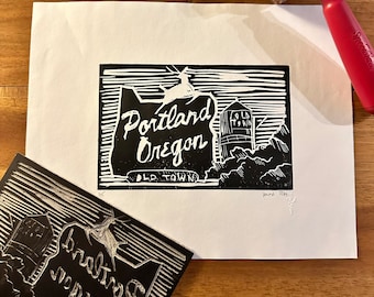 May include: A black and white linocut print of the Portland, Oregon skyline. The print features the iconic water tower and the text "Portland Oregon" with the words "Old Town" underneath. The print is signed and dated "June 2023".