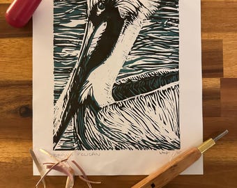May include: A linocut print of a brown pelican, with black, white, and teal ink. The print is on white paper, with the words "BROWN PELICAN" written in pencil. Printmaking tools, including a brayer and carving tools, surround the print.