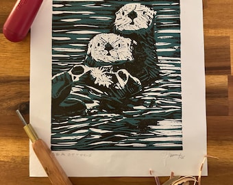 May include: A linocut print of two sea otters floating in water. The otters are black and white, with the water depicted in teal. The print is on white paper, with the words "sea otters" printed at the bottom. A red handled roller and carving tools are in the image.