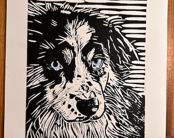 May include: Black and white linocut print of a dog's face with blue eyes. The dog's fur is detailed with white and black lines. The print is on a white paper with a wood grain background.
