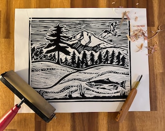 May include: Black and white linocut print depicting a mountain landscape with a pine tree, forest, and a flying bird. A salmon swims in the foreground. A red-handled brayer and a carving tool are on the wooden surface.