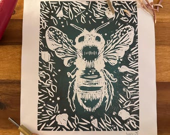 May include: A linocut print of a bee with a dark teal background and white details. The bee is surrounded by floral elements. Printmaking tools, including a brayer and carving tools, are visible on a wooden surface.