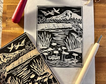 May include: A black and white linoleum block print of a mountain landscape with birds flying over a lake. The print is on a white sheet of paper. There are two wood carving tools and a brayer on a wooden surface.