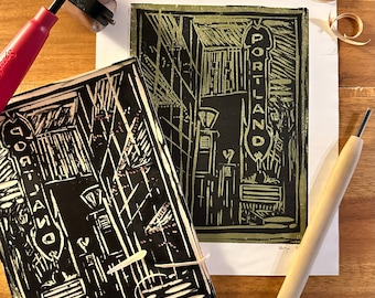 May include: Two black and white linoleum block prints of a city street scene with the word "PORTLAND" on a sign. The prints are on white paper and are surrounded by wood shavings and tools.