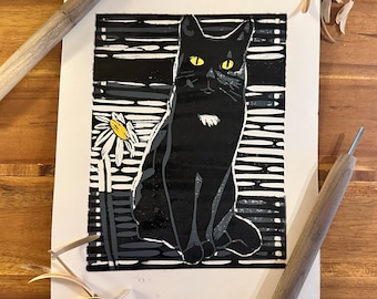 May include: A black and white linocut print of a cat with yellow eyes and a white patch on its chest. A daisy is to the left of the cat. Carving tools and wood shavings are around the print.