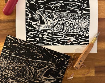 May include: A black and white linocut print of a fish with its mouth open, set against a watery background. A red handled brayer and a carving tool are visible. The print is on a white background, with a second print on a black background.