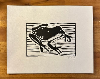 May include: Black and white linocut print of a frog. The frog is depicted in a side profile, with its legs extended. The print is on white paper and has a textured appearance. The print is signed and numbered 4/5.