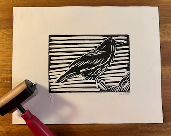 May include: Black and white linocut print of a raven perched on a branch, set against a striped background. The artwork is on white paper, with a red and tan printmaking roller in the foreground.
