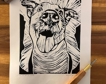 May include: Black and white linocut print of a dog with its tongue hanging out. The dog's face is the focal point, with detailed lines creating texture. The print is on white paper, with the word "ROCCO" visible.