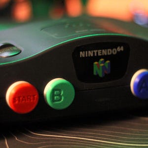 May include: Close-up of a black Nintendo 64 game console. The console features a red "START" button, a green "B" button, a blue "A" button, and a yellow button. The Nintendo 64 logo is visible on the console.