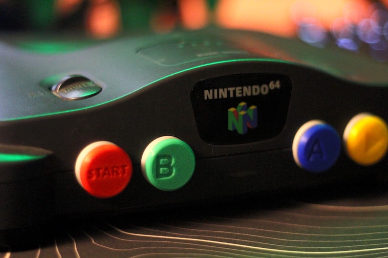 May include: Close-up of a black Nintendo 64 game console. The console features a red "START" button, a green "B" button, a blue "A" button, and a yellow button. The Nintendo 64 logo is visible on the console.