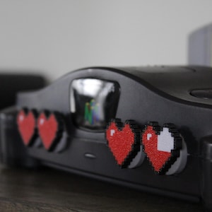 May include: A black Nintendo 64 console with pixelated red heart stickers. A cartridge with a Zelda graphic is visible in the background. The console is on a dark surface.