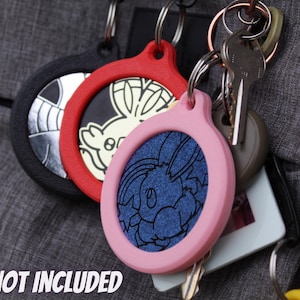 May include: Keychains in black, red, and pink, each with a circular design featuring a character illustration. The keychains are attached to a keyring with keys and a tag. The text "COIN NOT INCLUDED" is visible at the bottom.
