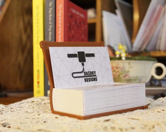Könnte beinhalten: Ein brauner Visitenkartenhalter in Buchform, gefüllt mit weißen Karten. Die oberste Karte zeigt ein geometrisches Muster und den Text "SHARKY DESIGNS". Er liegt auf einer Spitzendecke.
