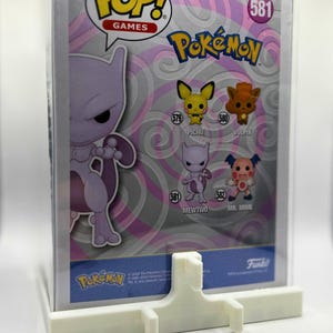 May include: A Funko Pop! Games Pokémon figure in a protective case, featuring Mewtwo and other characters. The packaging includes the Pop! Games logo and the number 581. A white pegboard stand is in front of the figure.