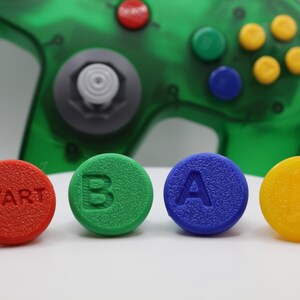 May include: A close-up of a green, translucent video game controller with four colorful buttons in the foreground. The buttons are red with "START", green with "B", blue with "A", and yellow. The controller has a gray joystick.