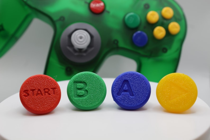 May include: A close-up of a green, translucent video game controller with four colorful buttons in the foreground. The buttons are red with "START", green with "B", blue with "A", and yellow. The controller has a gray joystick.