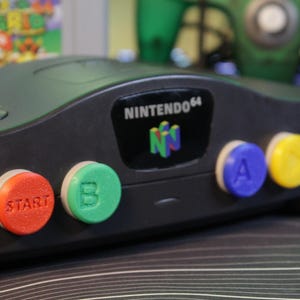 May include: A black Nintendo 64 game console with colorful buttons. The buttons are red, green, blue, and yellow. The console has the Nintendo 64 logo on it. A game cartridge is visible in the background.