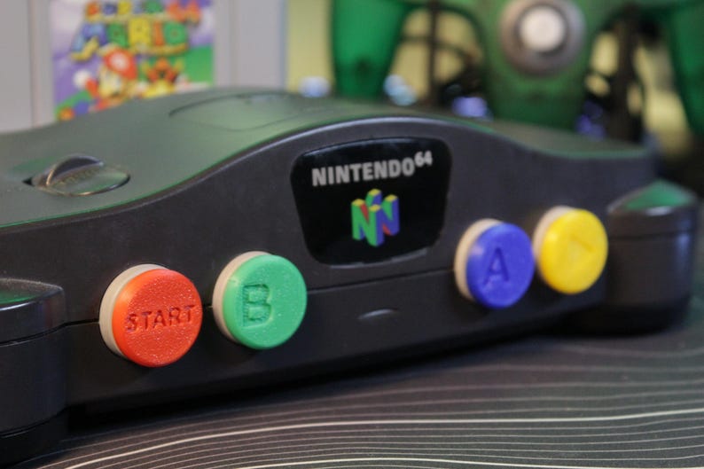 May include: A black Nintendo 64 game console with colorful buttons. The buttons are red, green, blue, and yellow. The console has the Nintendo 64 logo on it. A game cartridge is visible in the background.