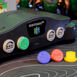 Puede incluir: Consola Nintendo 64 negra con botones coloridos. La consola tiene el logotipo de Nintendo 64 y varios botones verdes, blancos, azules, rojos y amarillos. Un cartucho de juego Super Mario 64 es visible en el fondo.