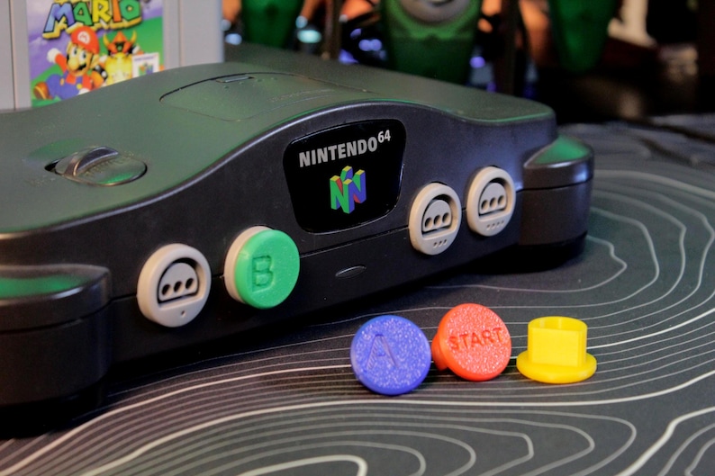 May include: Black Nintendo 64 console with colorful buttons. The console has the Nintendo 64 logo and several buttons in green, white, blue, red, and yellow. A Super Mario 64 game cartridge is visible in the background.