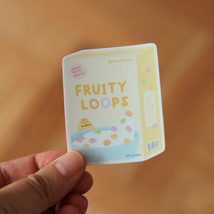 Puede incluir: Una pegatina de una caja de cereales de dibujos animados con un pollito amarillo en un tazón de bucles de cereales coloridos. La caja dice "Fruity Loops" en una fuente amarilla.